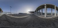 a spherical panoramic view shows some concrete structures and the sun behind them and a bus station