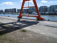 large red truss frame by the water with some docked boats in the background with people walking on walkway