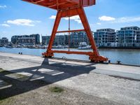 large red truss frame by the water with some docked boats in the background with people walking on walkway