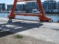 large red truss frame by the water with some docked boats in the background with people walking on walkway