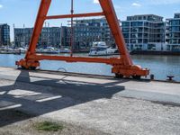 large red truss frame by the water with some docked boats in the background with people walking on walkway