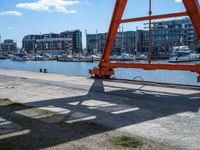 large red truss frame by the water with some docked boats in the background with people walking on walkway