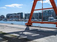 large red truss frame by the water with some docked boats in the background with people walking on walkway