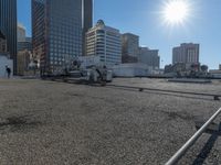 the truck is parked on the asphalt with the buildings in the background and a train going by