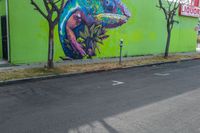 some graffiti artwork by two street signs along a green building and a tree in front