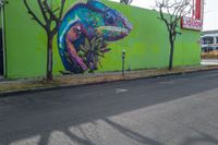 some graffiti artwork by two street signs along a green building and a tree in front