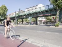 a woman riding her bicycle on the side of a road while a pedestrian crosses a bridge over the street