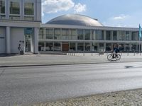 two people riding their bicycles past the building on the road, and pedestrians are outside