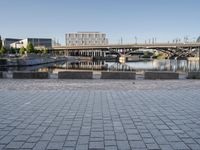 empty brick road near river with city skyline in the background at daytime, view of bridge, benches, and water