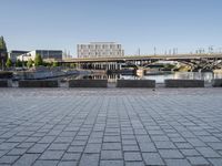 empty brick road near river with city skyline in the background at daytime, view of bridge, benches, and water