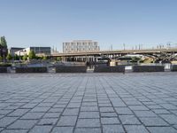 empty brick road near river with city skyline in the background at daytime, view of bridge, benches, and water