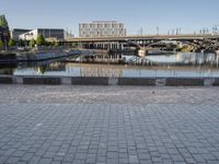 empty brick road near river with city skyline in the background at daytime, view of bridge, benches, and water