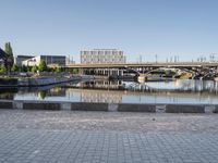 empty brick road near river with city skyline in the background at daytime, view of bridge, benches, and water