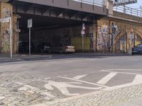 a paved road with bicycles and cars on the pavement beneath an underpass with graffiti