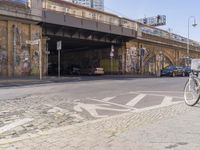 a paved road with bicycles and cars on the pavement beneath an underpass with graffiti