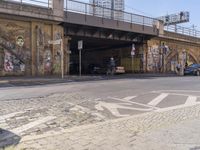 a paved road with bicycles and cars on the pavement beneath an underpass with graffiti