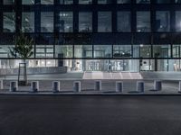 several concrete block benches and benches along a street at night near a building, there is lit up windows with reflective light