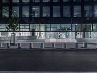 several concrete block benches and benches along a street at night near a building, there is lit up windows with reflective light