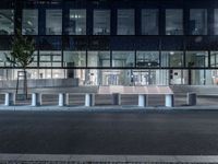 several concrete block benches and benches along a street at night near a building, there is lit up windows with reflective light