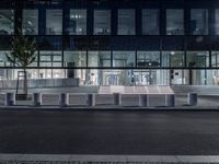 several concrete block benches and benches along a street at night near a building, there is lit up windows with reflective light