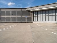 an empty parking lot has a no parking sign on the side of it and a large warehouse in the background