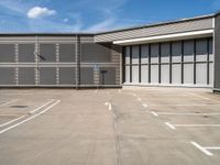 an empty parking lot has a no parking sign on the side of it and a large warehouse in the background