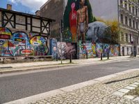 a wall with different colorful artwork on it in a city with buildings around and the street has a bike path going under it