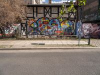 a wall with different colorful artwork on it in a city with buildings around and the street has a bike path going under it