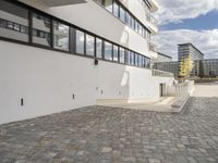 a concrete walkway leads to a building and other buildings in the background and blue skies with fluffy white clouds