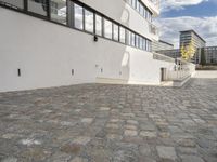 a concrete walkway leads to a building and other buildings in the background and blue skies with fluffy white clouds