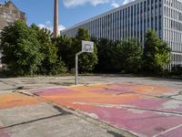 an abandoned basketball court with graffiti on it in a big building area with trees around