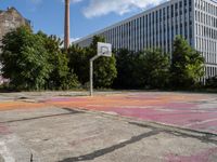 an abandoned basketball court with graffiti on it in a big building area with trees around