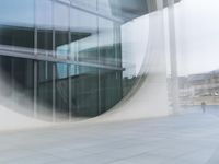 a man and his reflection on a round building window in the foreground, and a man with a helmet walking across an open courtyard