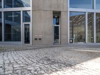 a grey brick sidewalk and some glass buildings and people standing outside of a building with people looking out the windows