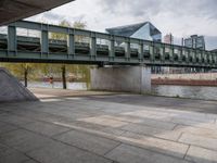 an image of city street under a green bridge in the day time with water and people walking around