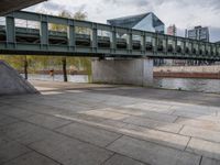 an image of city street under a green bridge in the day time with water and people walking around