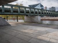 an image of city street under a green bridge in the day time with water and people walking around