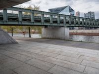 an image of city street under a green bridge in the day time with water and people walking around