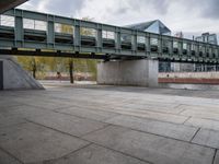 an image of city street under a green bridge in the day time with water and people walking around