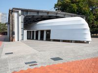 a white building with a roof in the middle of a sidewalk and a tree next to it