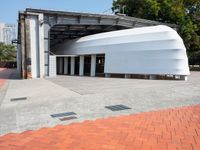 a white building with a roof in the middle of a sidewalk and a tree next to it