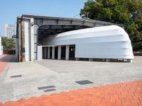 a white building with a roof in the middle of a sidewalk and a tree next to it