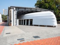 a white building with a roof in the middle of a sidewalk and a tree next to it