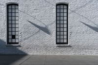 two windows on the white brick wall of an empty building in the shadows of another window