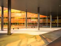 a long bench outside a building with glass windows on the side of the building and trees on the side of it