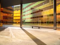 a large building is painted yellow and has glass walls and columns, with a bench outside of it