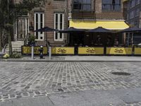 the sidewalk has two umbrellas near tables and benches outside it, and two people are sitting on it