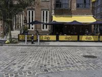 the sidewalk has two umbrellas near tables and benches outside it, and two people are sitting on it