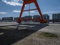 an orange crane is sitting in the field by some water and buildings on a sunny day