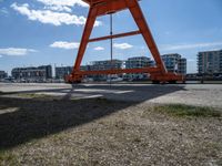 an orange crane is sitting in the field by some water and buildings on a sunny day